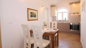 Ein Essbereich mit Holztisch und weißen Stühlen, eine Küche mit weißen Schränken und einem eingebauten Backofen sowie ein gerahmtes Bild an der Wand.