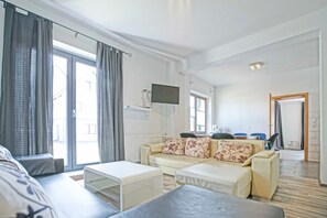 A modern living room with a sofa set, a flat-screen TV mounted on the wall, and a dining area with blue chairs.