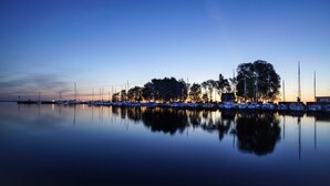 A marina at dusk with boats docked, calm water, and trees in the background.