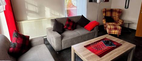 A living room with a grey sofa, a plaid armchair, a wooden coffee table, and a red and black patterned rug.
