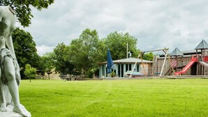 A statue in a grassy park area with a playground and a small building in the background. - Ferienhof - Weites - Land, large apartment with terrace, garden and children's playground (Stadtkyll)