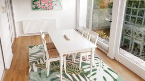 Un coin repas avec une table blanche, des chaises blanches, un tableau représentant des fleurs accroché au mur et une vue sur les arbres par la fenêtre.