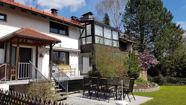 Une maison à deux étages avec un balcon, un petit jardin et une terrasse aménagée avec du mobilier d’extérieur.