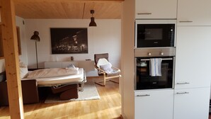 Un dormitorio con cama, mesita de noche, lámpara y una silla. Hay una zona de cocina con microondas, horno y armarios.