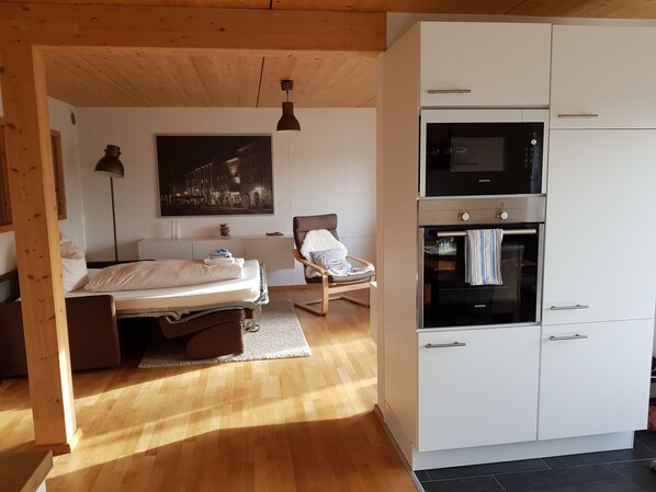 Un dormitorio con cama, mesita de noche, lámpara y una silla. Hay una zona de cocina con microondas, horno y armarios.