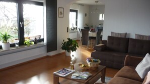 Een woonkamer met een bruine bank, een salontafel met tijdschriften, een potplant en een eethoek op de achtergrond.