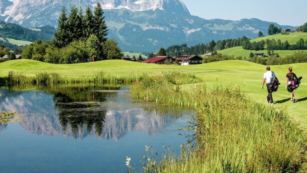 Ein Golfplatz mit Teich, Häusern und Bergen im Hintergrund.