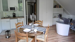 Eine moderne Küche mit weißen Schränken, einer Mikrowelle und einer Kaffeemaschine. Ein Esstisch mit Stühlen und einer Kerzenleuchter steht dort. Eine Treppe führt zu einer Schlafgalerie mit einer Couch und Bücherregalen.