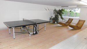 Ein Tischtennis-Tisch, ein Holzboden, ein Fenster mit Blick auf Grünflächen und ein Holzstuhl.