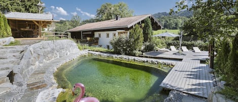 A rustic chalet with a wooden deck, a swimming pool with a pink flamingo float, and a stone pathway.
