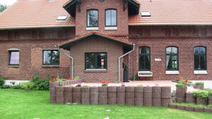 A two-story brick house with a red-tiled roof, multiple windows, and a front garden with flowers and rocks. - Large farmhouse with pool, minutes from the beach  (Greifswald)