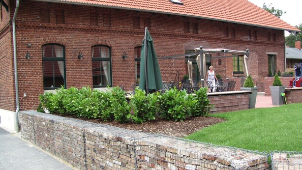 A brick building with a red-tiled roof, a small patio area with a table and chairs, and a green umbrella. - Large farmhouse with pool, minutes from the beach  (Greifswald)