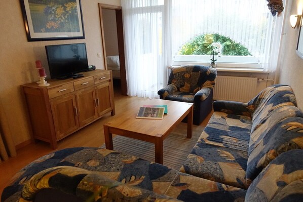 A living room with a sofa, a wooden coffee table, a television on a cabinet, and a window with a view of greenery.