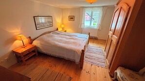 Een slaapkamer met een houten bed, nachtkastjes, een nachtkastje, een raam met gordijnen, een houten garderobe en een plafondlamp.