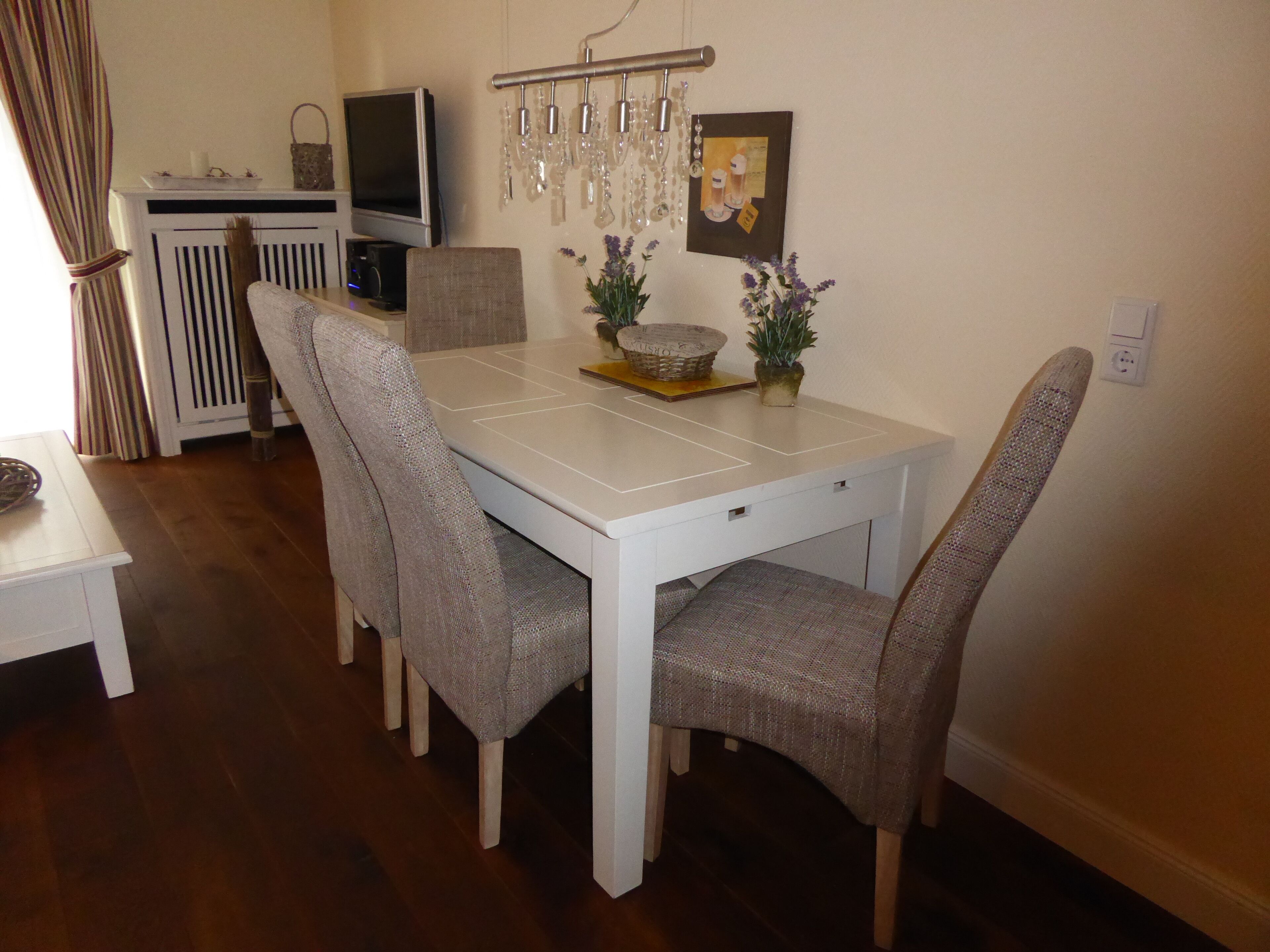 A dining area with a white table, four upholstered chairs, a television, a chandelier, and a framed picture on the wall.