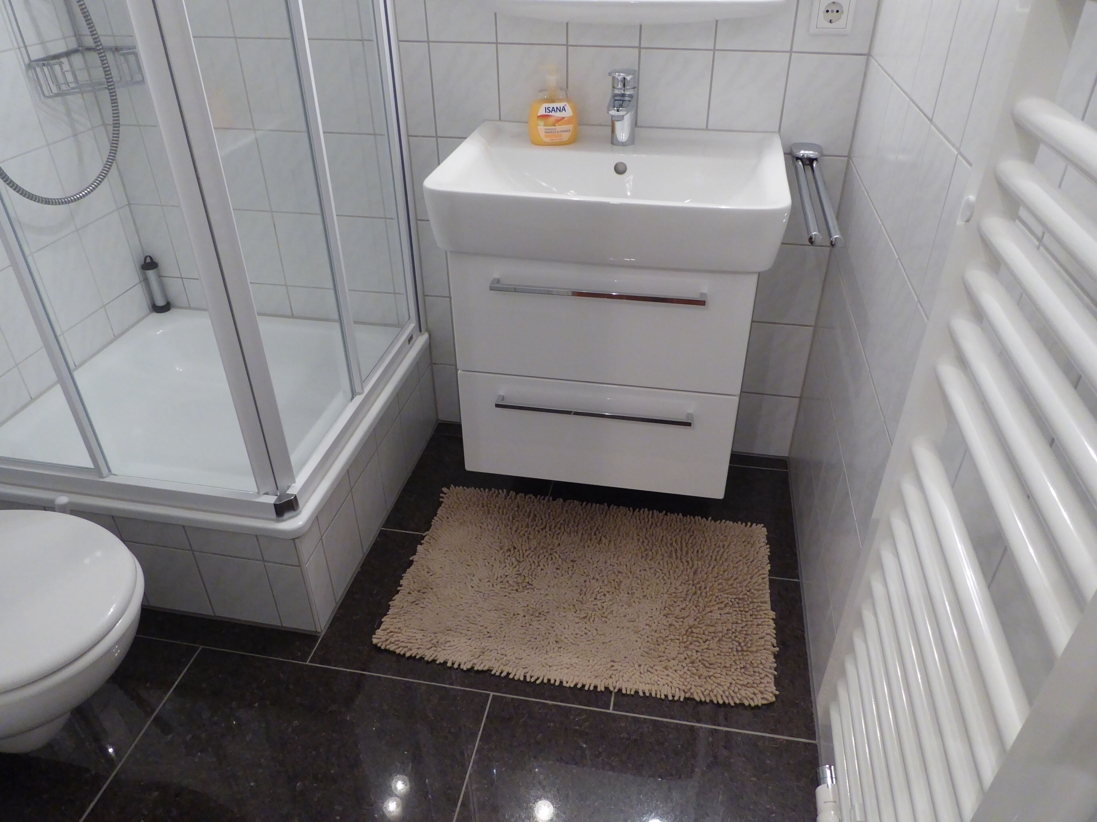 A bathroom with a glass-enclosed shower, a white sink with a soap dispenser, and a beige rug on the floor.