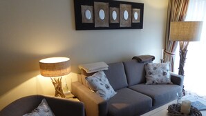 Uma sala de estar com sofá, poltrona e mesa de centro. Há um abajur sobre uma mesinha lateral e uma obra de arte na parede, acima do sofá.