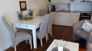 Uma sala de jantar com uma mesa branca e cadeiras cinzentas, uma cozinha com armários brancos e um fogão, e uma sala de estar com um sofá cinza e um copo decorativo sobre uma mesa branca.