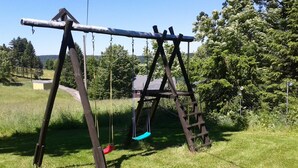 A wooden swing set in a grassy field with trees and a house in the background.