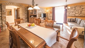 A dining area with a long wooden table, chairs, and a fruit bowl. There is a stone wall and a television in the background. - Private and secure 5000 sqm garden, pool, children's playground, only 9 km from the sea (Nedešćina)