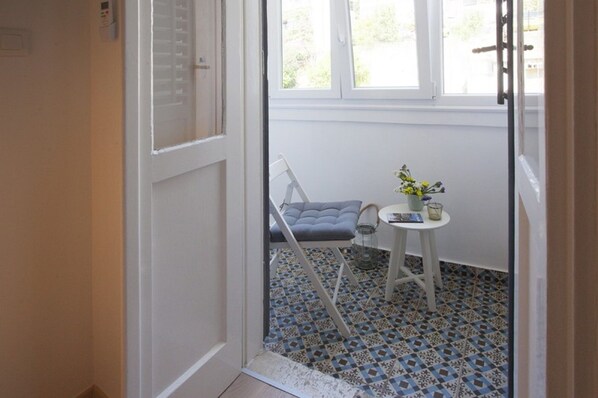A small room with a white door, a blue patterned floor, a grey cushioned chair, a small round table with a vase of flowers, and a window with a view of greenery. - Studio apartment Xenia Split****, waterfront promenade, old town, air conditioning, loggia (Split)