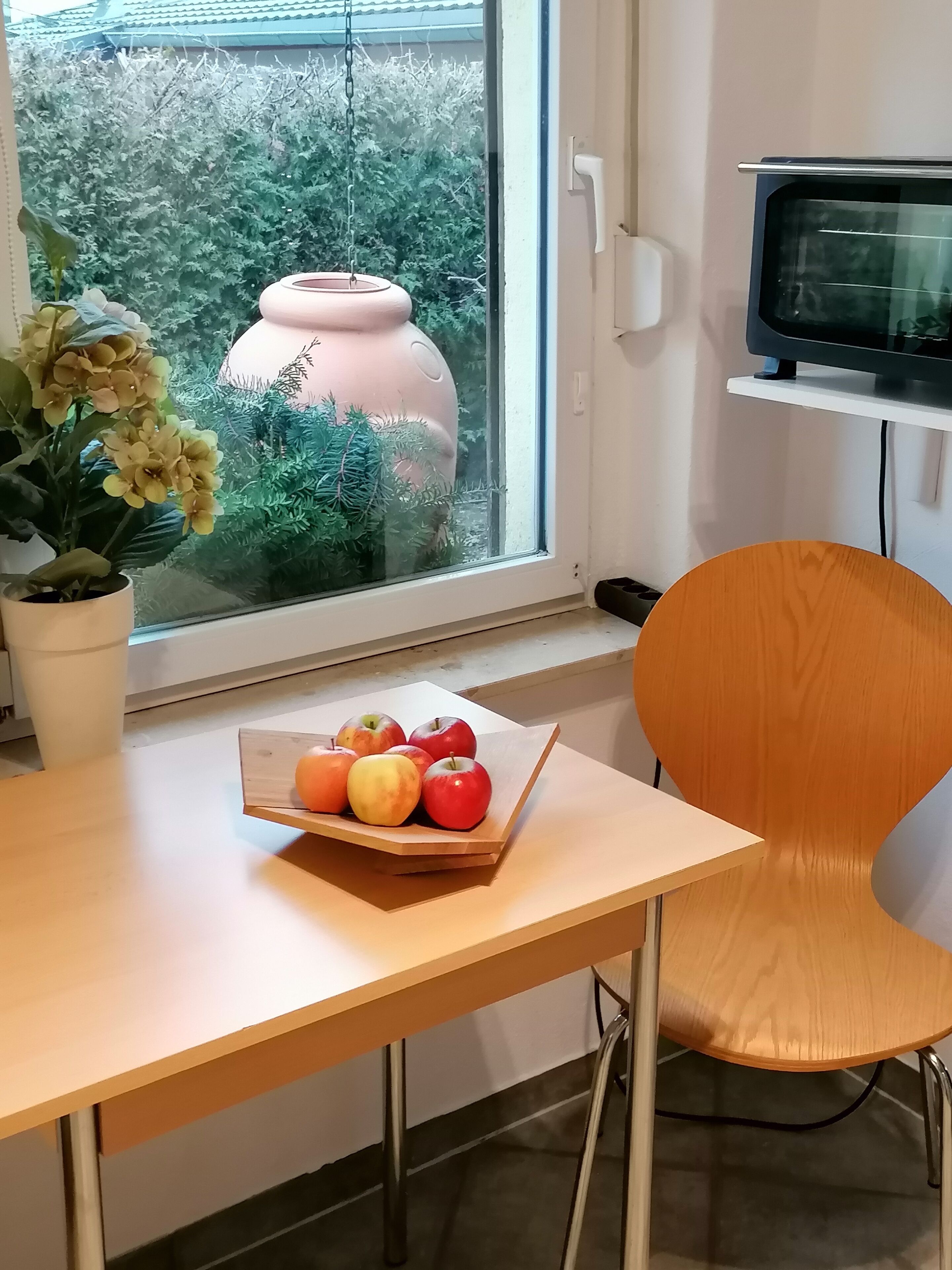 Una habitación con ventana, una planta en maceta, una mesa con frutero y un televisor.