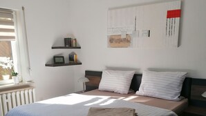 A modern bedroom with a large bed, a bedside table, a wall-mounted shelf with books, and a window with curtains.