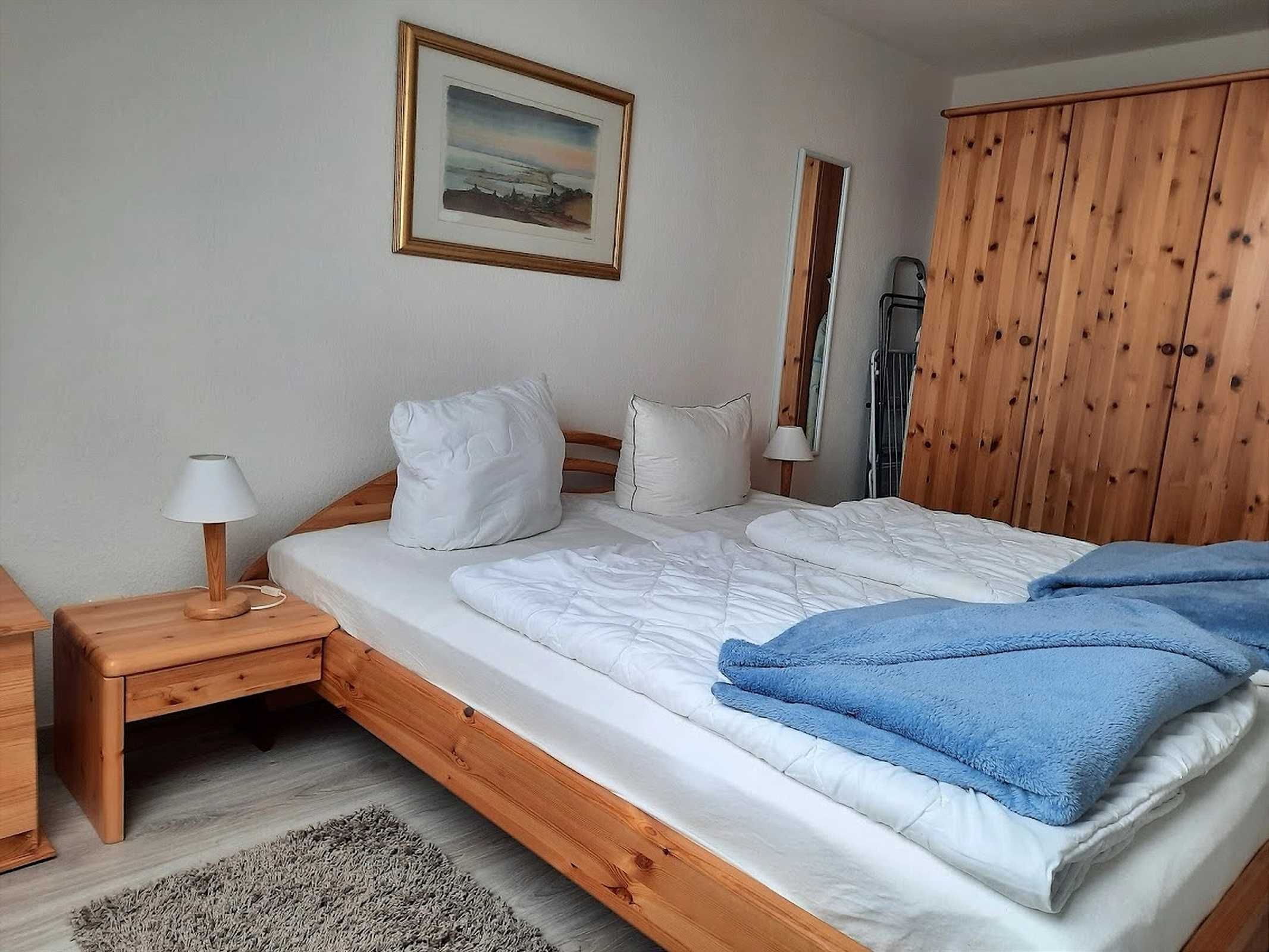 A bedroom with a wooden bed, white bedding, a blue blanket, a bedside lamp, and a framed picture on the wall.