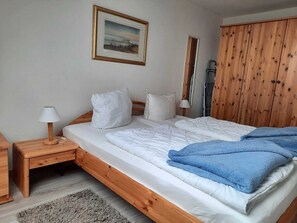 Un dormitorio con una cama de madera, ropa de cama blanca, una manta azul, una lámpara de noche y un cuadro en la pared.