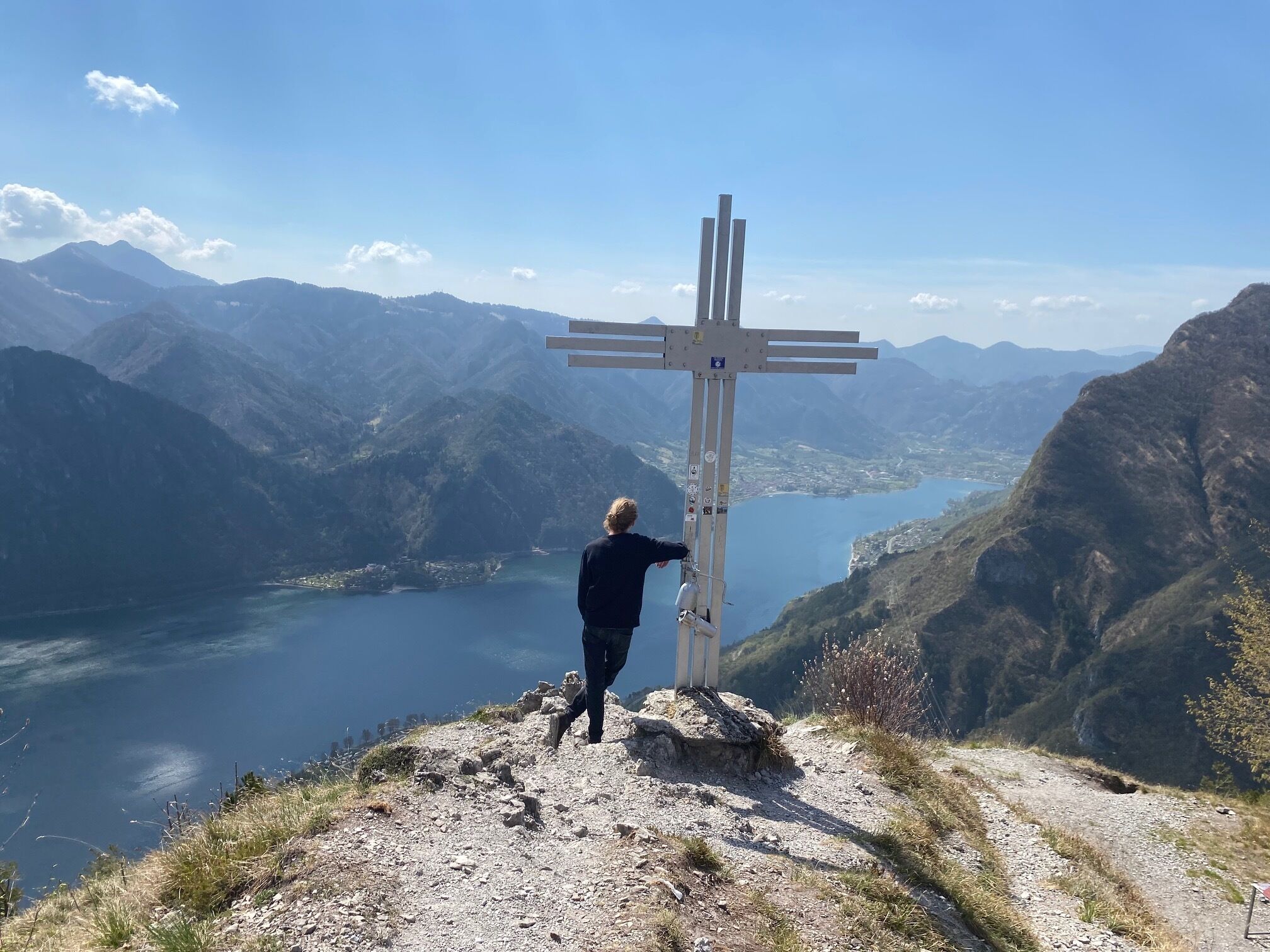 Henkilö seisoo vuoren huipulla olevan ristin vieressä, ja taustalla näkyy järvi ja vuoristo.