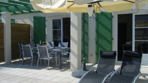 Eine Außenpatio mit Tisch und Stühlen, ein überdachter Sitzbereich mit Sonnenschirmen und ein Gebäude mit grünen Fensterläden.