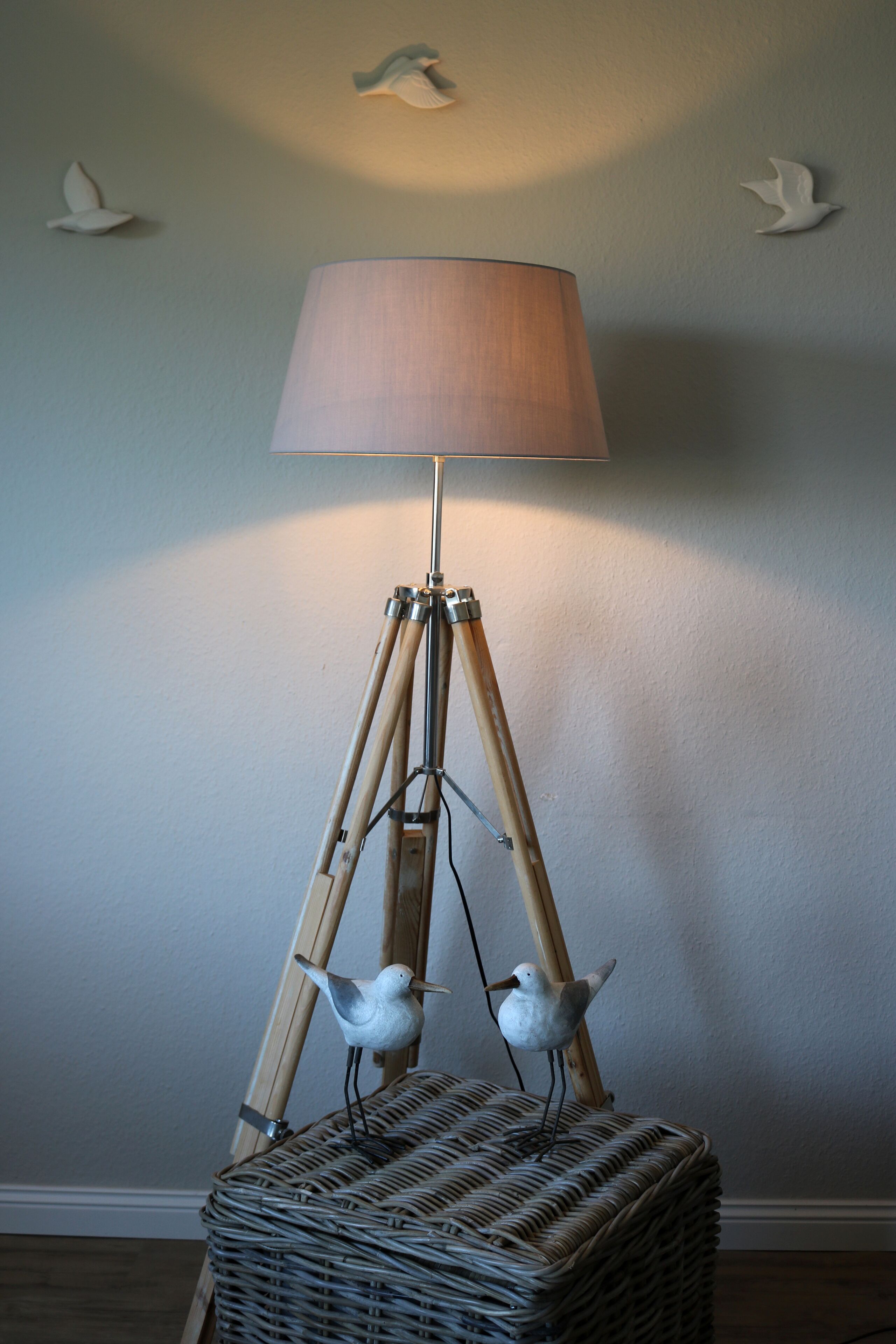Une lampe de sol dotée d’un abat-jour beige, montée sur un pied tripode, placée à côté d’un panier en osier.