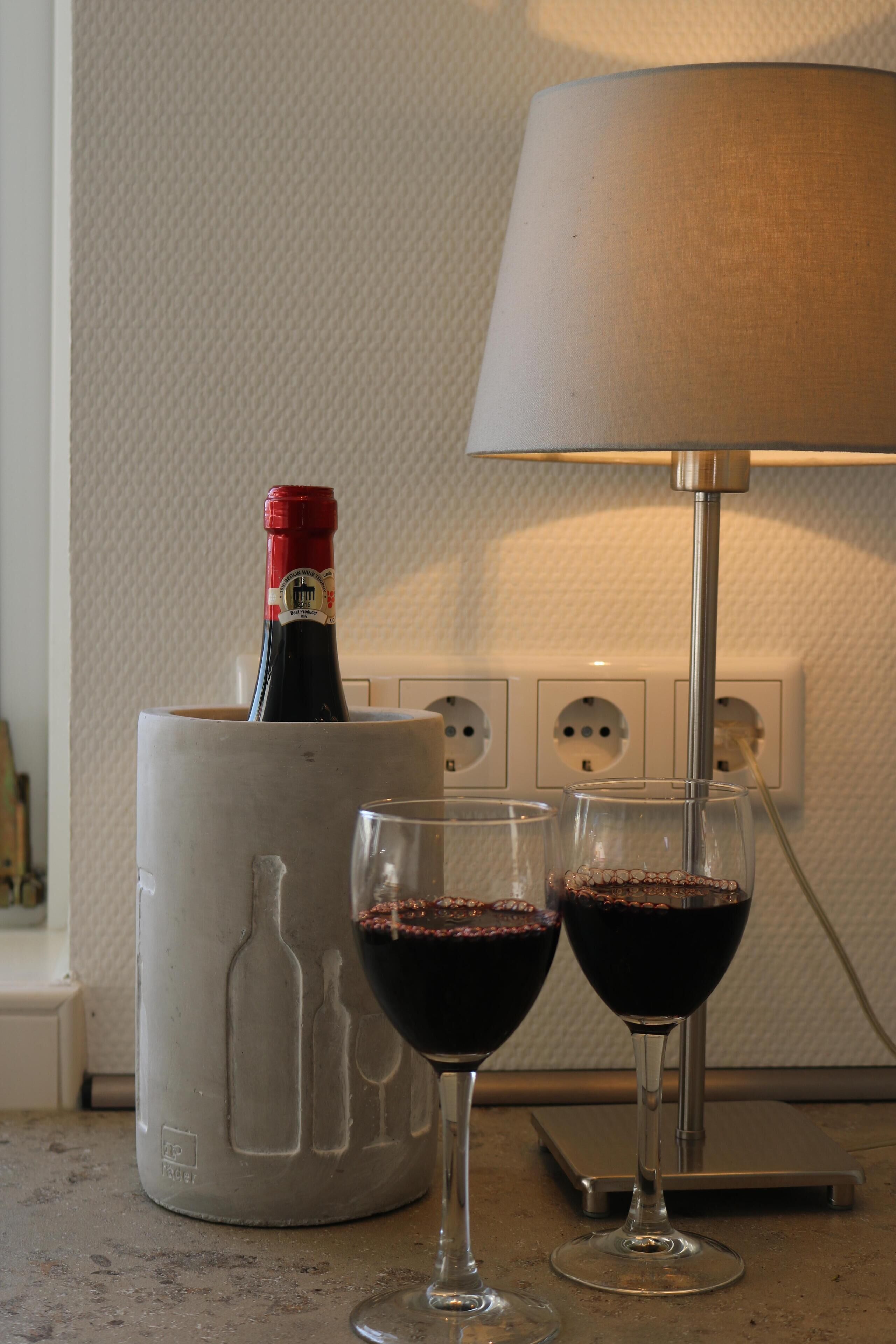 Una botella de vino tinto en un recipiente decorativo, dos copas de vino llenas de vino tinto y una lámpara de mesa sobre una mesa.
