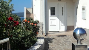 A stone pathway leading to a white building with a tiled roof, a metal gate, and a bush with red flowers.