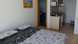 A compact bedroom with a bed, pillows, and a painting on the wall. A small kitchenette is visible in the adjacent room.