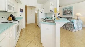 Une cuisine compacte équipée d’un four à micro-ondes, d’une cuisinière et d’un réfrigérateur. On y trouve un évier avec un robinet et un plan de travail avec un égouttoir. La cuisine a un sol carrelé et une porte qui donne sur une autre pièce.