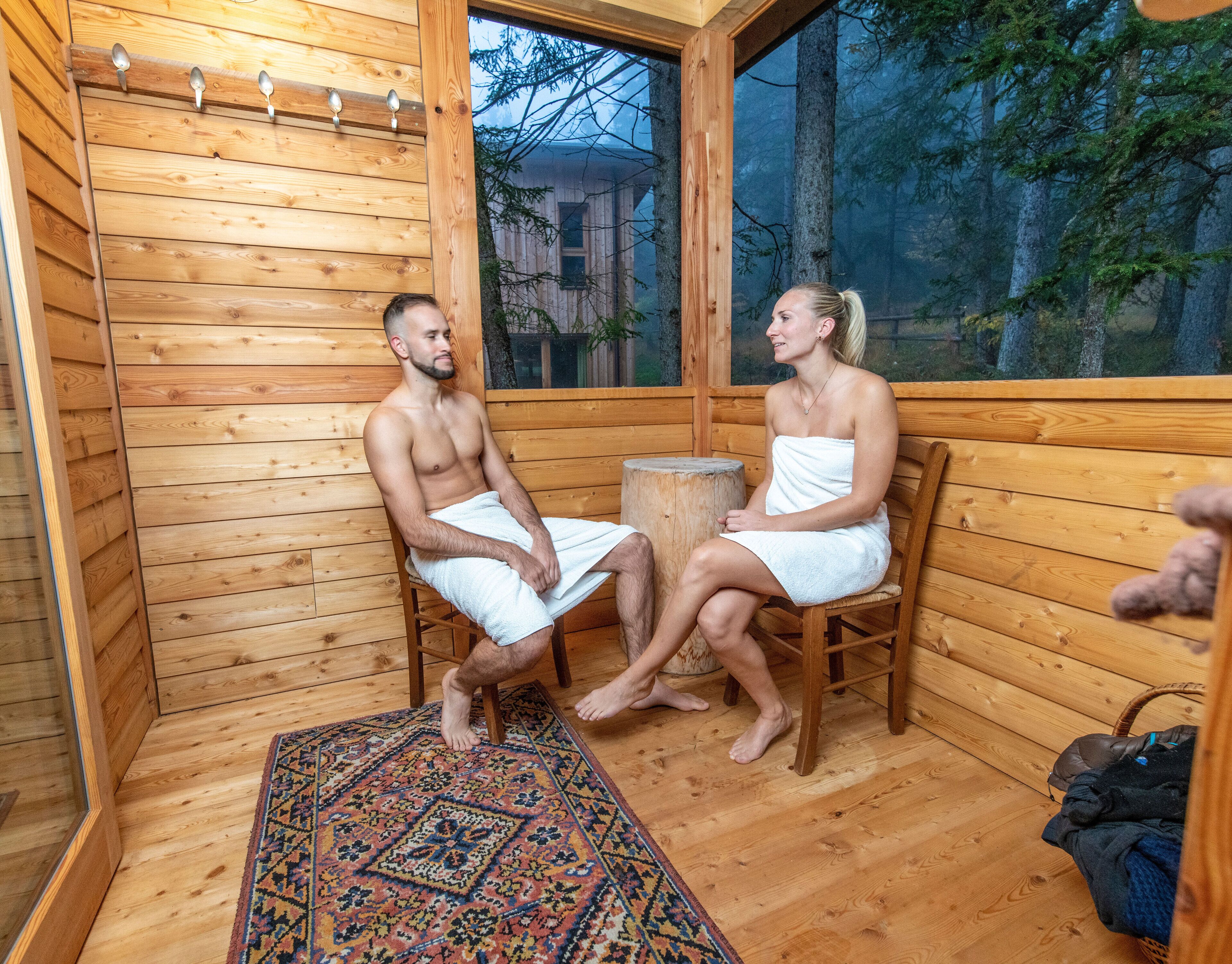 A wooden sauna room with a large window, a wooden bench, and a rug on the floor.