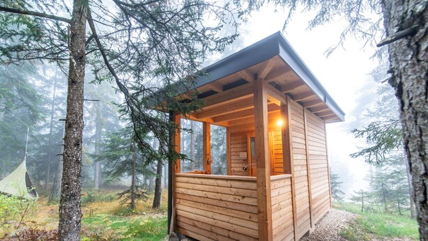 A wooden cabin with a sloped roof, large windows, and a visible interior light.