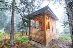 A wooden cabin with a sloped roof, large windows, and a visible interior light.