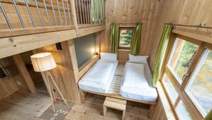 A wooden cabin with a loft bed, two single beds, a small table, and a window with curtains.