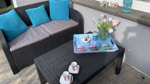 Een rieten bank met teal kussens, een klein tafeltje met een dienblad waarop een vaas met roze bloemen staat, een teal lantaarn en een raam met gordijnen.