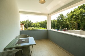 Een balkon met een glazen tafel en rieten stoelen, met uitzicht op bomen en een gebouw op de achtergrond.