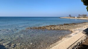 Une plage côtière aux eaux claires et peu profondes, avec un rivage rocheux et quelques bâtiments visibles au loin.