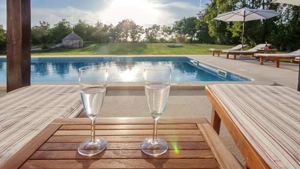 Une table en bois, avec deux verres d’eau, donnant sur une piscine bordée de chaises longues et de parasols.
