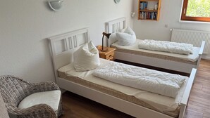 Et værelse med to hvide senge, en fletstol, et natbord med lampe, en bogreol og et vindue med udsigt til grøn bevoksning.