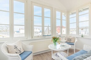 Ein helles Zimmer mit großen Fenstern, Korbmöbeln und einem Glastisch mit Blumen.