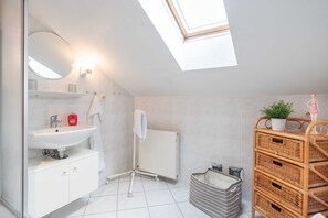 A modern bathroom with a skylight, white tiles, a white sink, a white radiator, and a wicker storage unit. - Apartment 3 - HausSeebär (Heringsdorf)