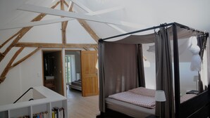 A bedroom with a four-poster bed, wooden beams, a bookshelf, and a bathroom visible through an open door.