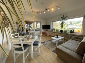 A modern living room with a dining area, a sofa, and a large window with a view of houses.