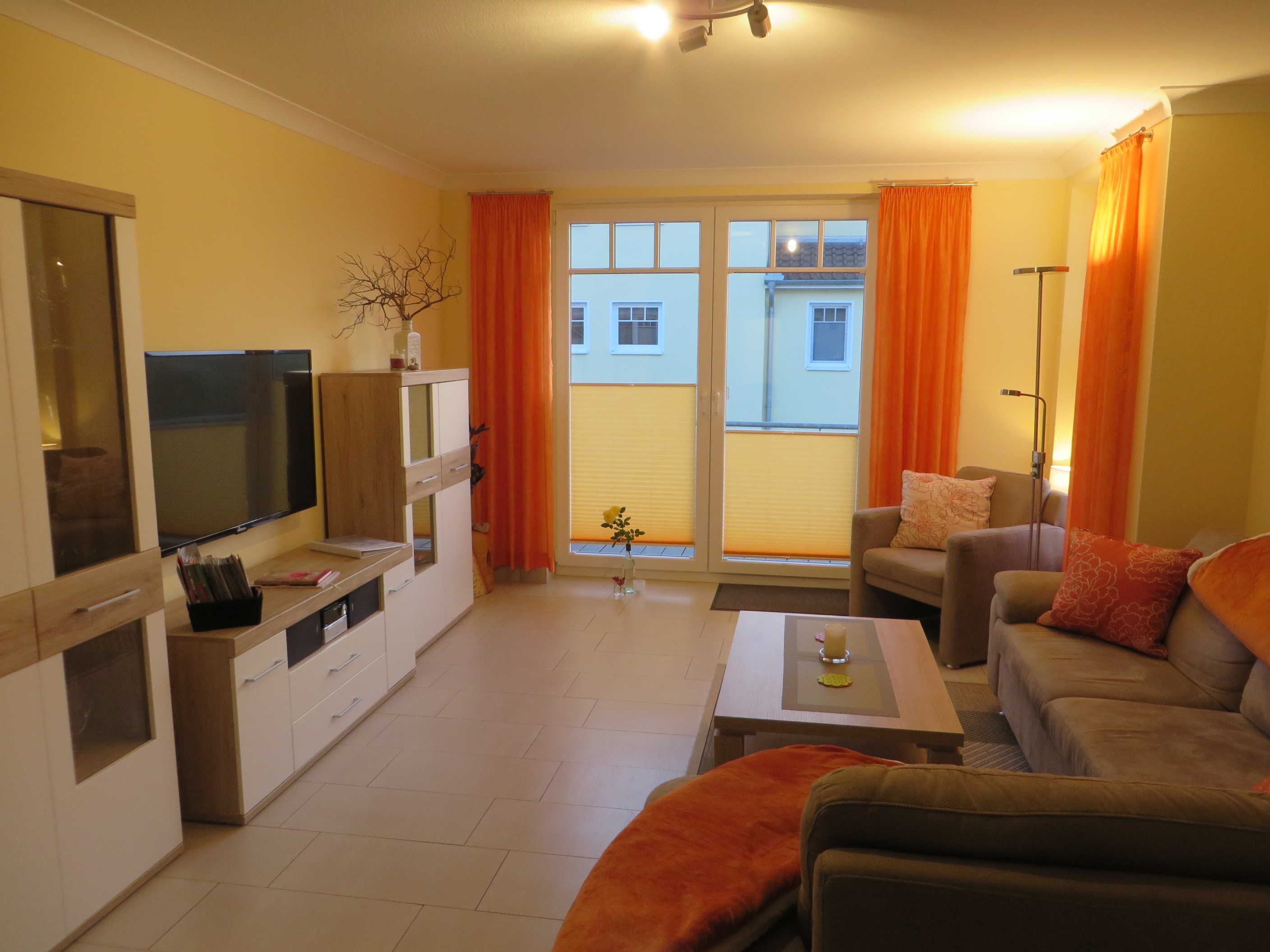 Ein Wohnzimmer mit einer Couch, einem Couchtisch und einem Fernseher. Ein Balkon mit orangenen Vorhängen und Blick auf Gebäude.